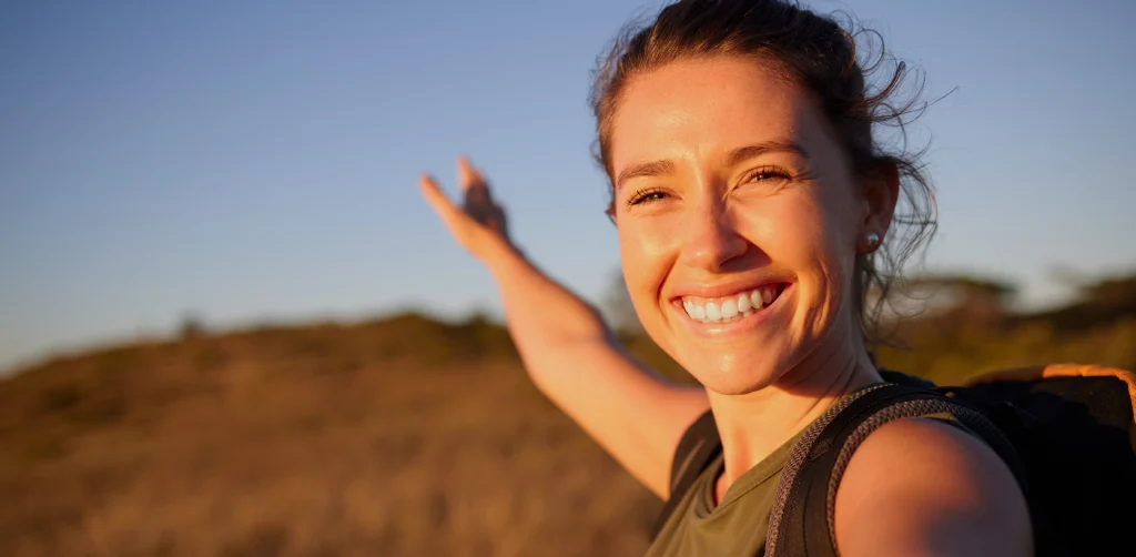 A woman smiling in warm evening light on a hillside walk, hand raised